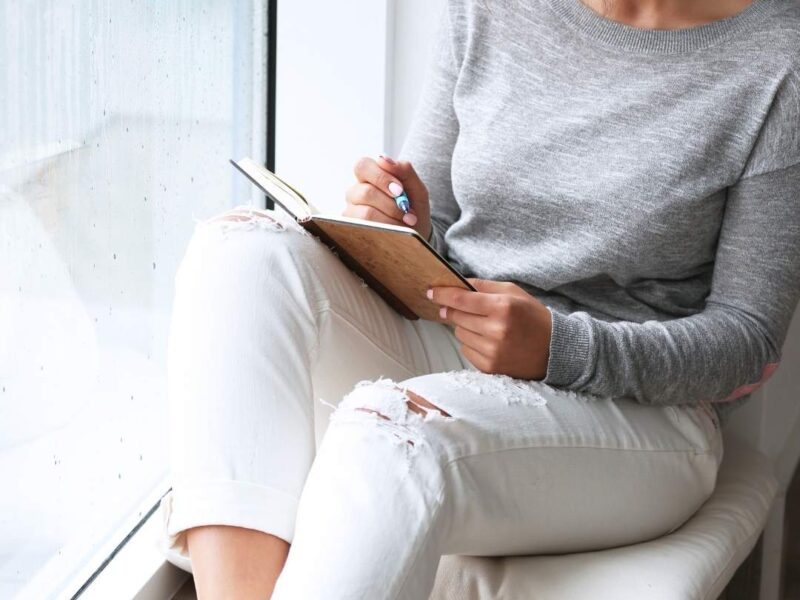 A-featured-image-of-self-care-goals-for-the-new-year-showing-a-woman-sitting-at-the-windoq-journaling-her-new-year-goals