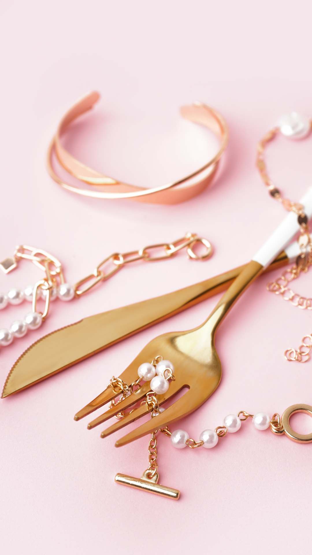 Minimalistic-jewelries-for-simple-and-everyday-pieces-including-a-twisted-cuff-bracelet-chain-necklace-and-pearl-accents-artfully-arranged-with-fork-and-knife-on-a-pink-background