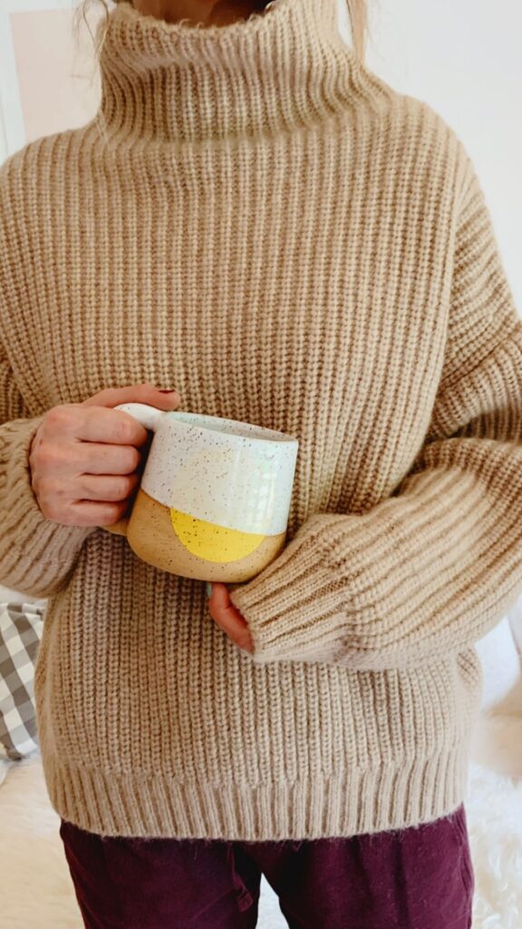 A-woman-wearing-light-brown-cozy-sweater-while-holding-a-brown-and-white-cup-as-her-layered-fall-outfits