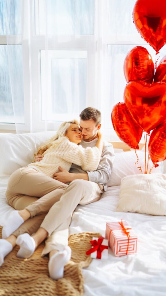 Cozy-indoor-setup-for-couples-valentines-day-photoshoot-ideas-showing-lovers-cuddled-up-wearing-cream-colored-outfits-with-ared-balloon-and-gift-box-beside-them